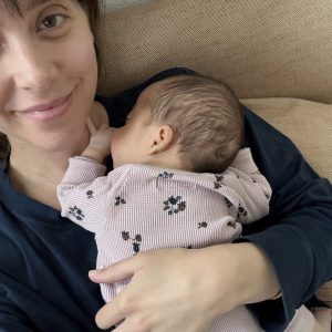 A woman smiles gently while holding a sleeping baby in her arms. The baby wears a floral-patterned outfit.
