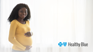 Pregnant woman smiling gently, holding her belly, in a bright room with soft curtains. "Healthy Blue" logo is in the bottom right corner.