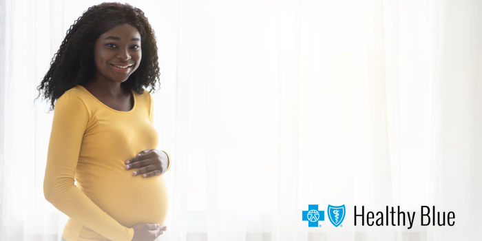 Pregnant woman smiling gently, holding her belly, in a bright room with soft curtains. "Healthy Blue" logo is in the bottom right corner.