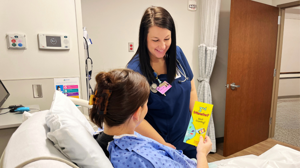 A nurse in blue scrubs smiles while handing a Count the Kicks brochure to a patient in a hospital bed. The atmosphere is warm and caring.